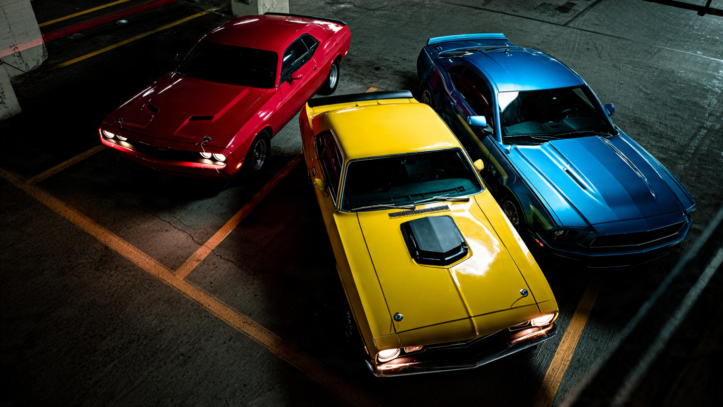 Three American muscle cars parked side by side in a dimly lit garage: a red Dodge Challenger, a yellow Chevrolet Camaro, and a blue Ford Mustang